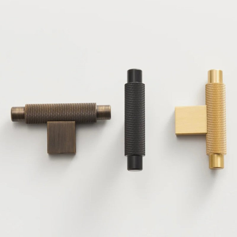 A brass cabinet pull with an antique black and gold finish features a cross-shaped T-bar design and a knurled texture for grip.